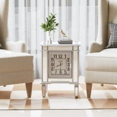 A stylish vintage clock on a white side table between two cream armchairs, decorated with a glass vase of green leaves and a bird figurine, in a bright room with sheer curtains; perfect for modern or classic home decor.