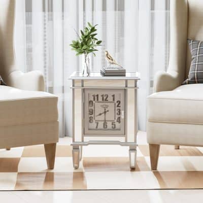 A stylish vintage clock on a white side table between two cream armchairs, decorated with a glass vase of green leaves and a bird figurine, in a bright room with sheer curtains; perfect for modern or classic home decor.