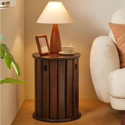 A stylish wooden bedroom side table with a round top, featuring a contemporary design. It is placed beside a white sofa with beige cushions, holding a table lamp, a framed photo, and a cup, enhancing a warm and inviting home ambiance. Suitable for modern interior decor, this side table adds functional elegance to living or bedroom spaces.