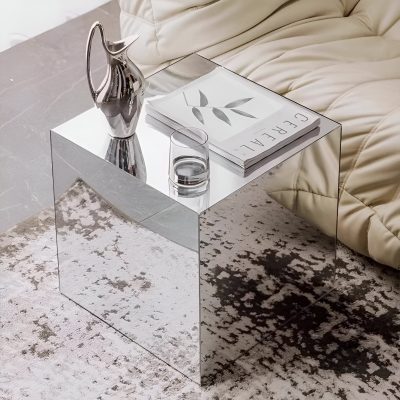 A sleek, reflective glass mirror coffee table placed beside a beige sofa, decorated with a water jug, glass, and a magazine, offering a contemporary and stylish accent for living room decor.