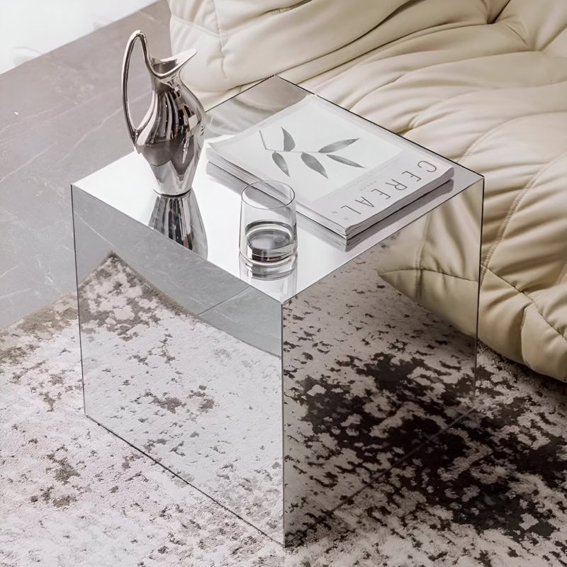 A sleek, reflective glass mirror coffee table placed beside a beige sofa, decorated with a water jug, glass, and a magazine, offering a contemporary and stylish accent for living room decor.