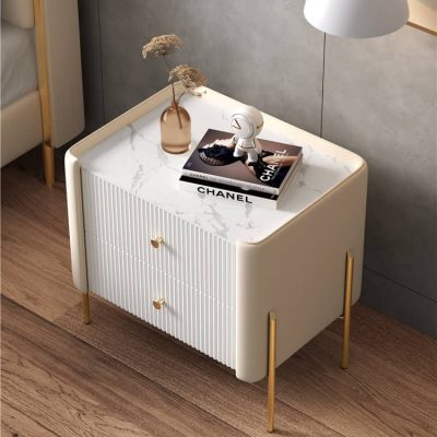 A stylish white nightstand featuring a marble top, gold legs, and dual drawers, perfect for modern bedroom decor. Includes a Chanel book, decorative vase with dried flowers, and a small lamp.