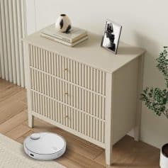 A white bedroom dresser with slatted doors, gold knobs, and minimalist design sits beside a robotic vacuum cleaner on wooden flooring. Decorated with contemporary accessories, it offers functional storage and enhances bedroom aesthetics.