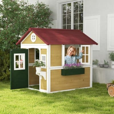 Bright yellow and white children's playhouse with a red roof, featuring a smiling young girl peeking out the window, surrounded by greenery and potted plants in a backyard setting.