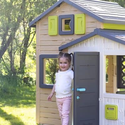 Cute young girl smiling at the camera while standing at the door of a colorful outdoor playhouse in a sunny, green backyard.