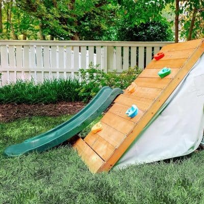 Slide climbing wall with plastic holds and a green slide in a backyard garden.
