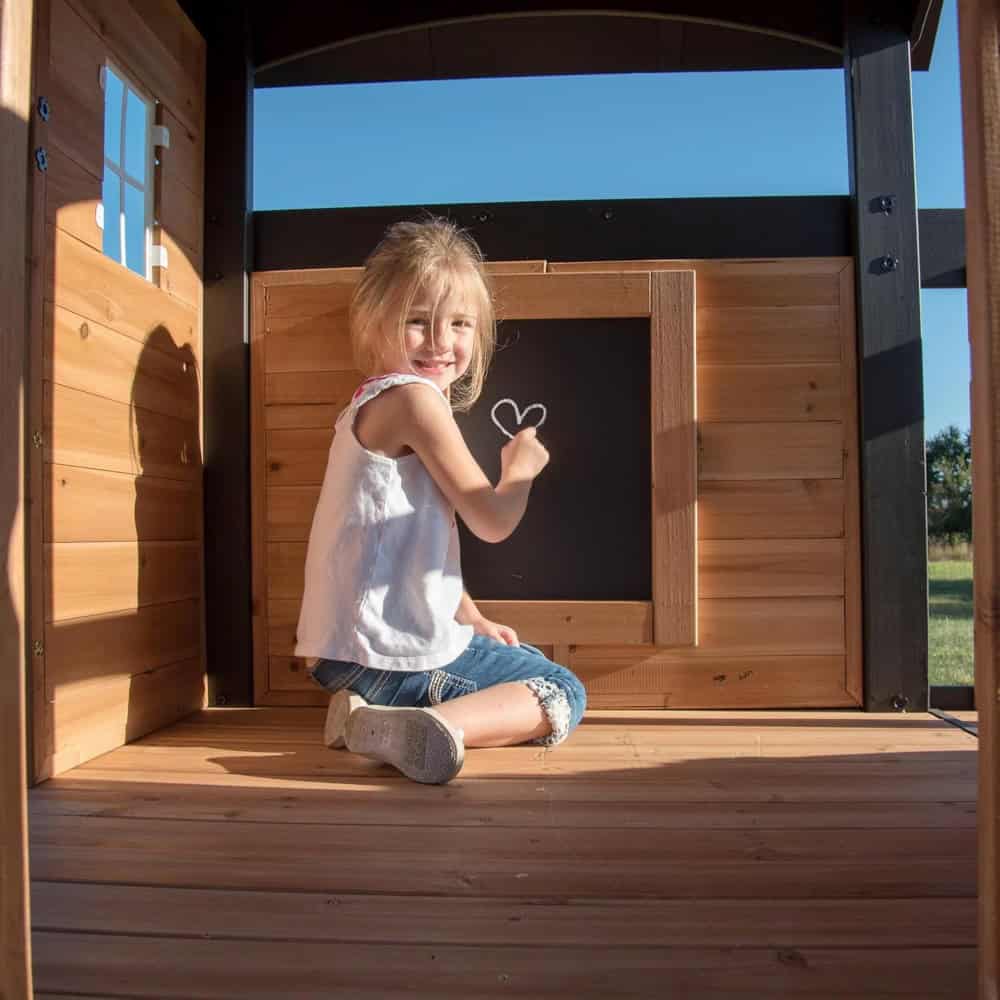 Child drawing heart on blackboard in wooden outdoor playhouse, emphasizing children’s toys and outdoor activities at Monalisa Store.