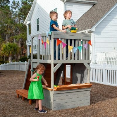 Children playing on a suburban backyard wooden playhouse with sandpit, colorful flags, and children enjoying outdoor fun.
