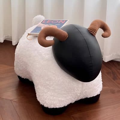 Fluffy sheep-shaped ottoman with black body and brown curled horns, placed on a wooden floor, next to a white curtain, with a magazine on top.