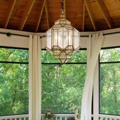 Elegant geometric chandelier hanging indoors with wooden ceiling and large windows showing lush greenery outside.