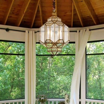 Elegant geometric chandelier hanging indoors with wooden ceiling and large windows showing lush greenery outside.