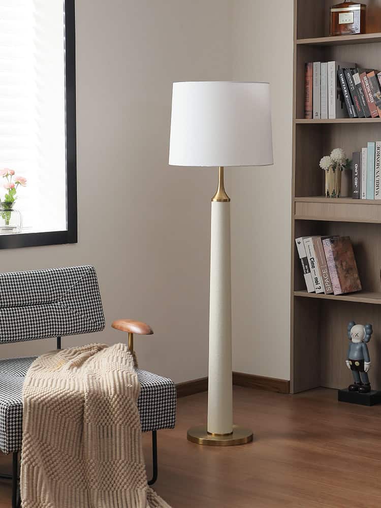 Modern floor lamp with white shade and gold accents in a cozy living space featuring a black and white houndstooth armchair, wooden bookshelf with books and decorative items, and natural light from a nearby window.