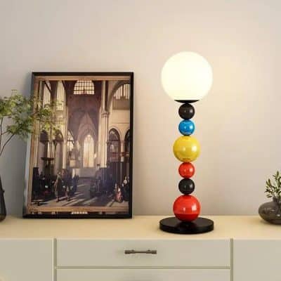 A modern table lamp with a colorful stacked ball base and a glowing round white shade, placed on a white dresser with decorative items and a framed building interior photo in the background.
