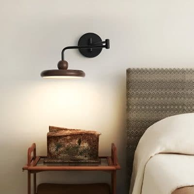 Contemporary wall-mounted wooden reading lamp in black and brown design, illuminating a cozy bedroom corner with a textured headboard and vintage art piece on a small wooden table.