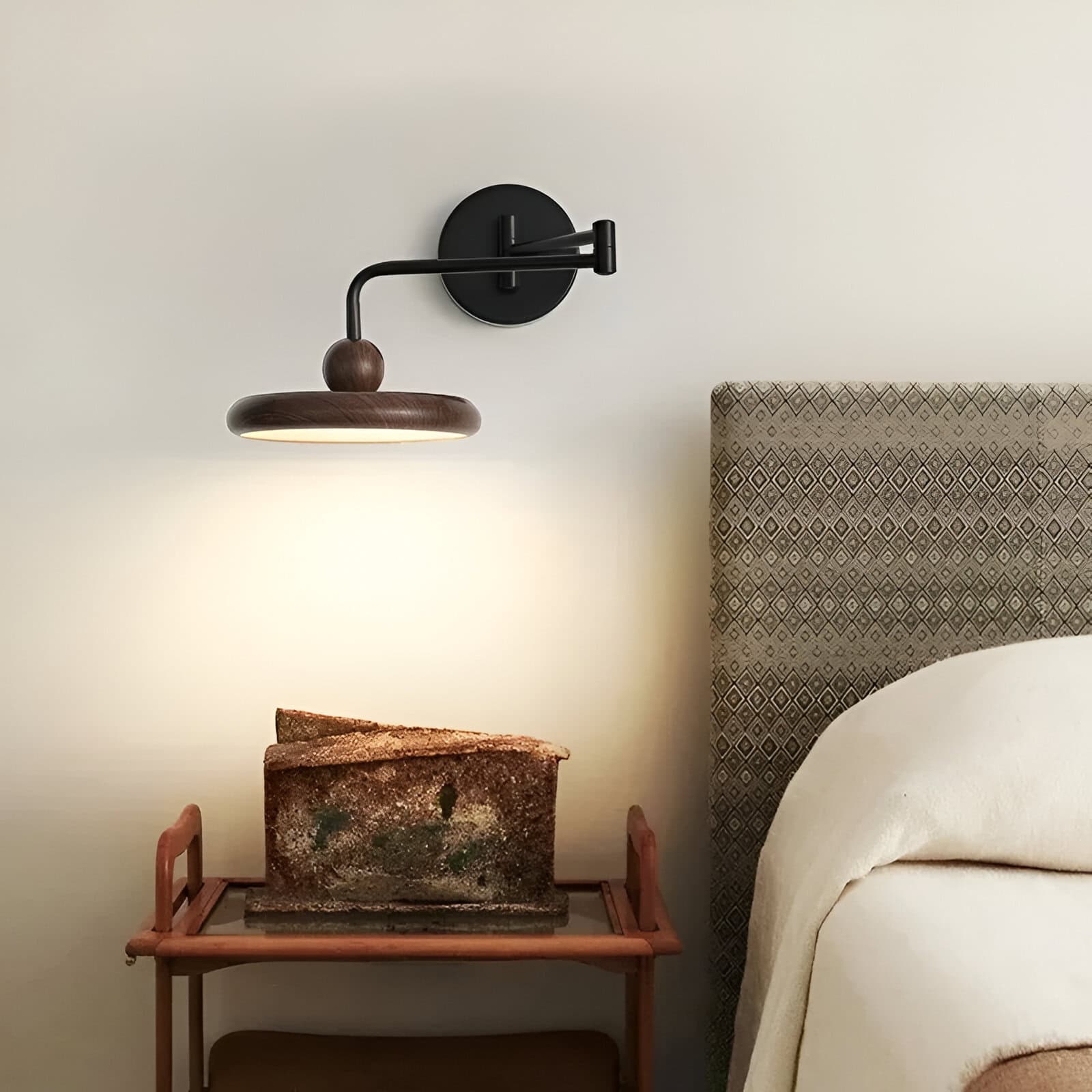 Contemporary wall-mounted wooden reading lamp in black and brown design, illuminating a cozy bedroom corner with a textured headboard and vintage art piece on a small wooden table.