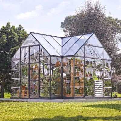 Greenhouse with glass panels filled with tropical plants, set outdoors with trees and blue sky in the background. Suitable for gardening and plant growth, showcasing modern greenhouse design at Monalisa Store.