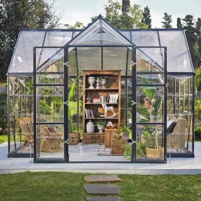 Stylish glass greenhouse with black metal frame, lush greenery, wooden furniture, and decorative items, perfect for garden enthusiasts and outdoor living enthusiasts.