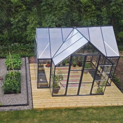 Modern glass greenhouse with black frame, solar panels, and wooden deck in a lush garden setting. Ideal for gardening enthusiasts seeking a stylish, functional outdoor green space.