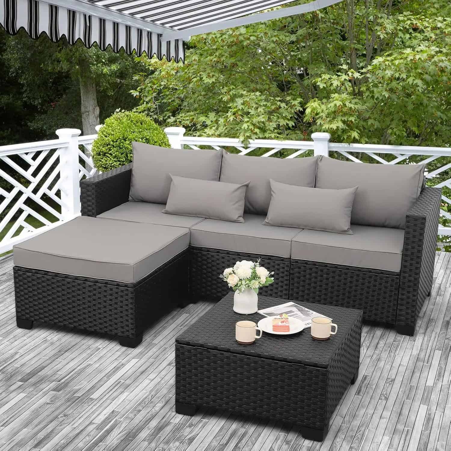 Outdoor patio furniture set with dark wicker design, cushions, and a small table with coffee mugs, flowers, and magazines, perfect for relaxing or entertaining in a garden or backyard setting.