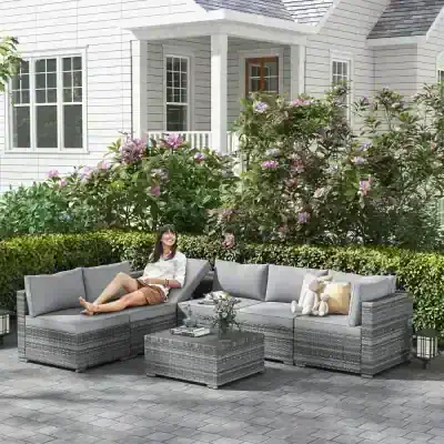 Elegant outdoor patio furniture set with a gray wicker sectional sofa, coffee table, and cushions, perfect for relaxing in a beautifully landscaped garden with blooming pink flowers.