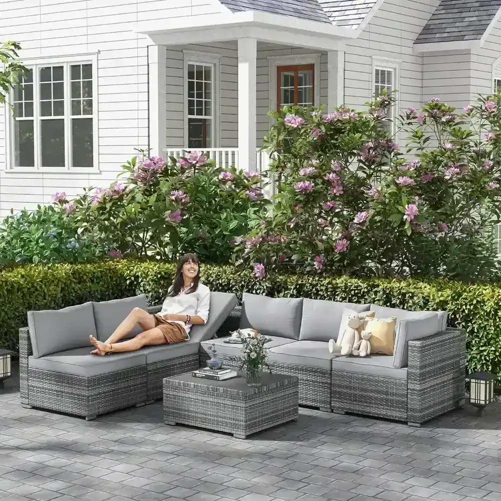 Elegant outdoor patio furniture set with a gray wicker sectional sofa, coffee table, and cushions, perfect for relaxing in a beautifully landscaped garden with blooming pink flowers.