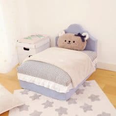 Cozy children's bed with a plush bear pillow, soft blanket, striped bedsheet, and bedside storage, ideal for a nursery or kids' bedroom.