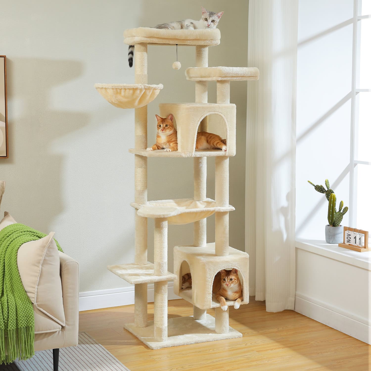 Fluffy beige cat tree with multiple perches, hideouts, and lounging platforms adorned with three cats, placed in a bright living room with natural sunlight, curtains, and a potted cactus.