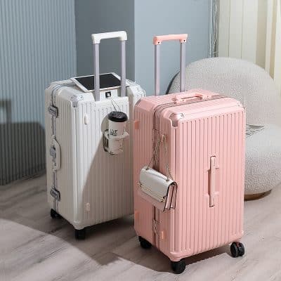 White and pink rolling luggage with telescoping handles and a small handbag attached, placed in a modern waiting area with a plush chair and light-colored flooring.