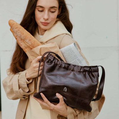 A woman holding a bouquet of bread and a newspaper, carrying a black leather bag, with the Monalisa Store logo visible on the bag, showcasing stylish accessories and daily essentials.