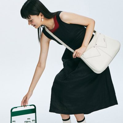 Vibrant woman dressed in a fashionable black skirt and red shoes, carrying a white handbag, reaching for bottled water on a miniature green trolley, embodying trendy style and everyday elegance.