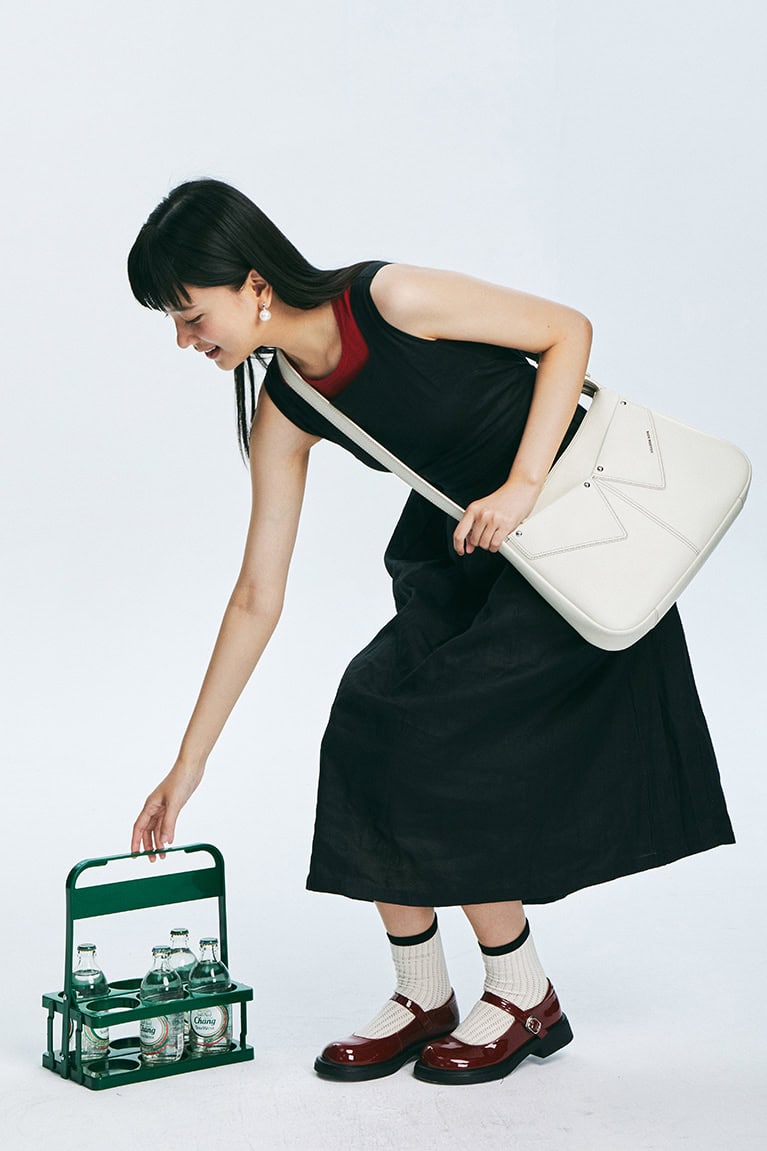 Vibrant woman dressed in a fashionable black skirt and red shoes, carrying a white handbag, reaching for bottled water on a miniature green trolley, embodying trendy style and everyday elegance.