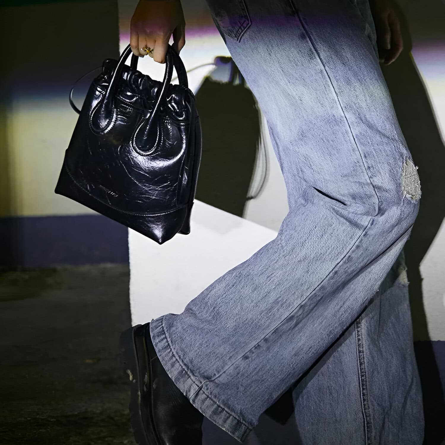 Black leather handbag held by person wearing distressed denim jeans and black boots, in a modern, stylish setting.