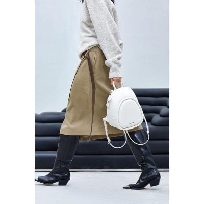 Stylish woman holding a white backpack, wearing a beige skirt and black boots, in modern indoor setting. Perfect for fashion-forward accessories and trendy outfit ideas.