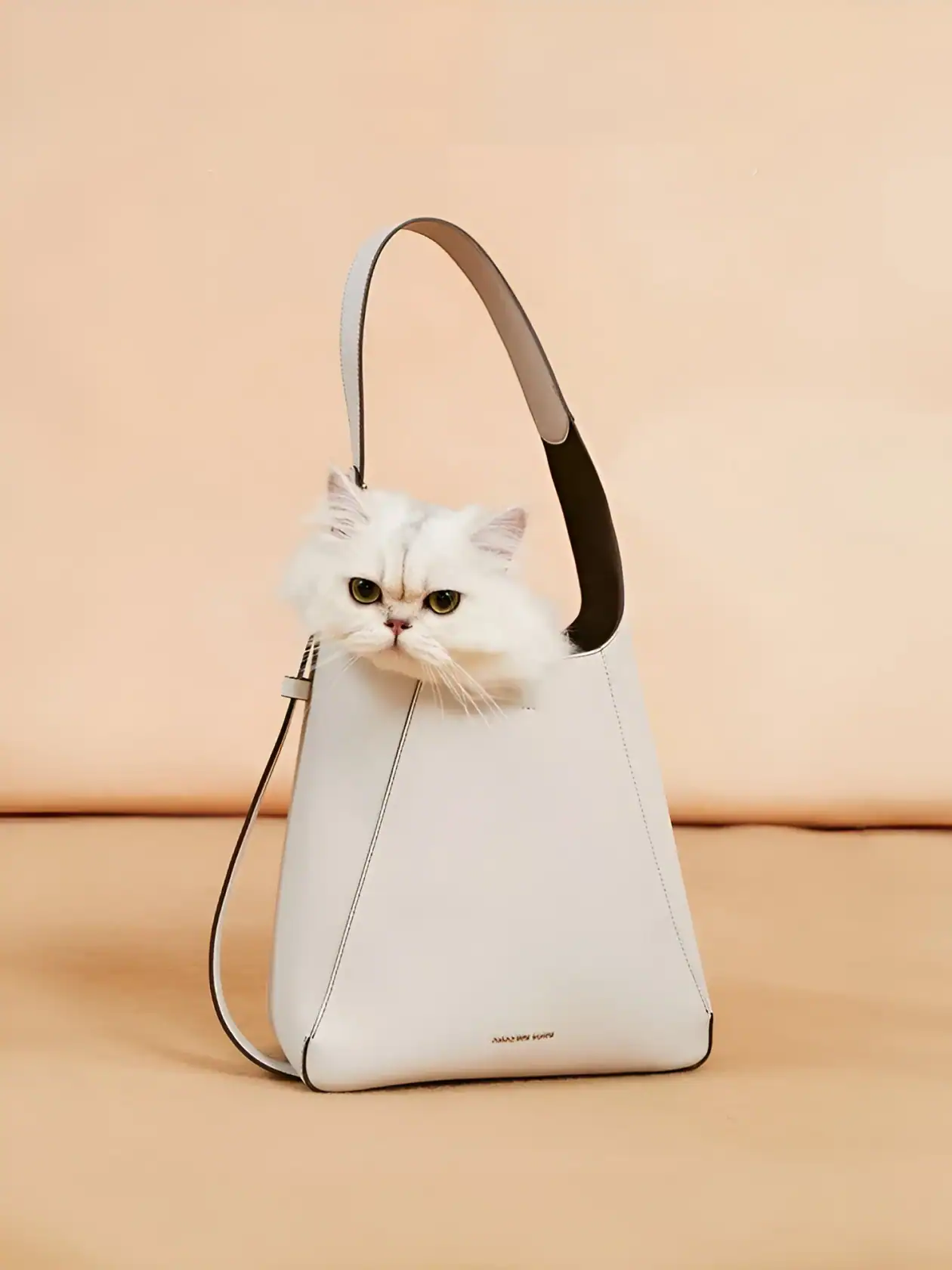 Fluffy white Persian cat with yellow eyes peeking out of a sleek, modern white handbag on a peach background, showcasing luxury pet accessories from Monalisa Store.