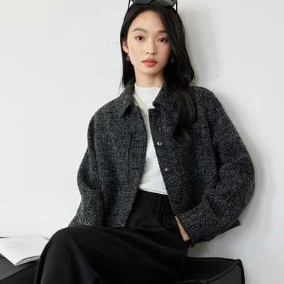Stylish young woman wearing a black textured jacket and black wide-leg pants, sitting against a white wall with sunglasses on her head, showcasing modern fashion and beauty style at Monalisa Store.
