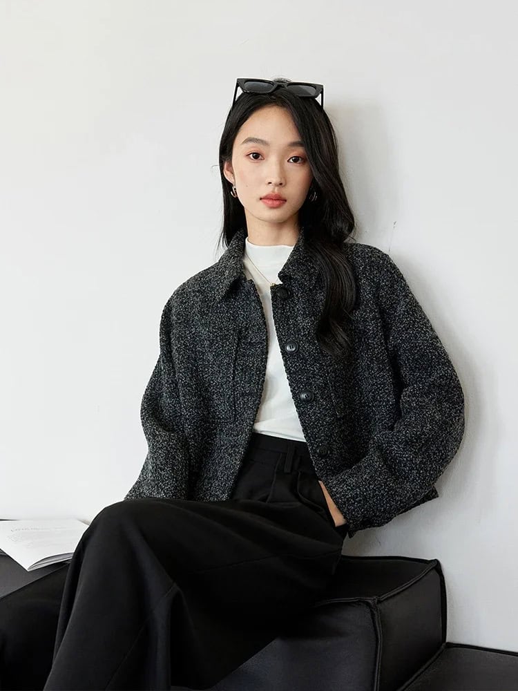 Stylish young woman wearing a black textured jacket and black wide-leg pants, sitting against a white wall with sunglasses on her head, showcasing modern fashion and beauty style at Monalisa Store.