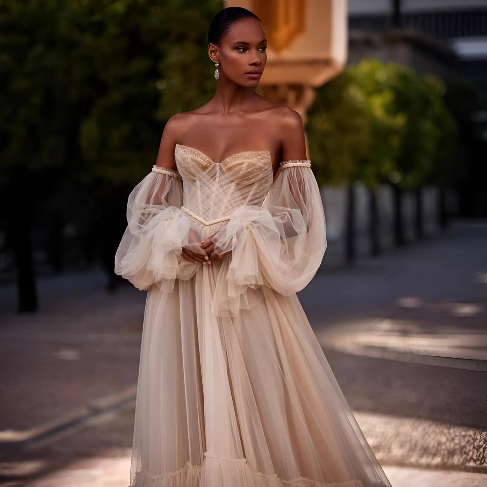 Elegant woman in a beige evening gown with sheer puff sleeves, standing outdoors during sunset, showcasing luxury fashion and sophisticated style.