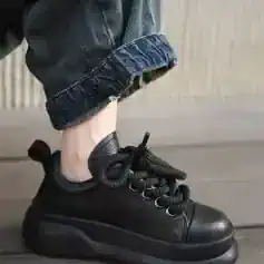 Black chunky fashion sneaker with thick sole, textured leather, and unique knotted laces; worn with rolled-up denim jeans.