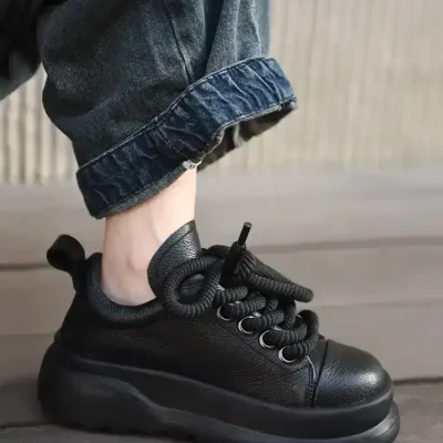 Black chunky fashion sneaker with thick sole, textured leather, and unique knotted laces; worn with rolled-up denim jeans.