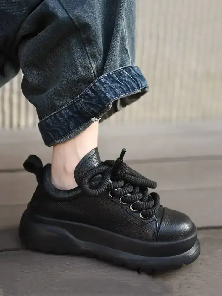 Black chunky fashion sneaker with thick sole, textured leather, and unique knotted laces; worn with rolled-up denim jeans.
