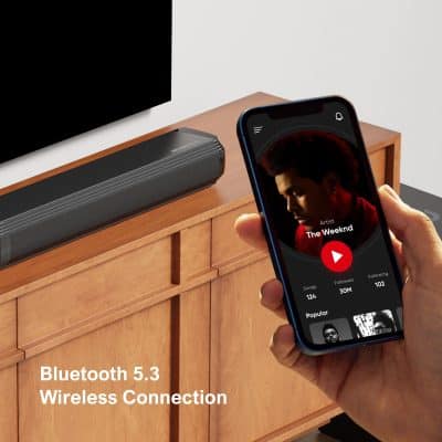 High-quality Bluetooth 5.3 wireless speaker on wooden cabinet, with a person's hand holding a smartphone showing music app, showcasing modern audio technology.