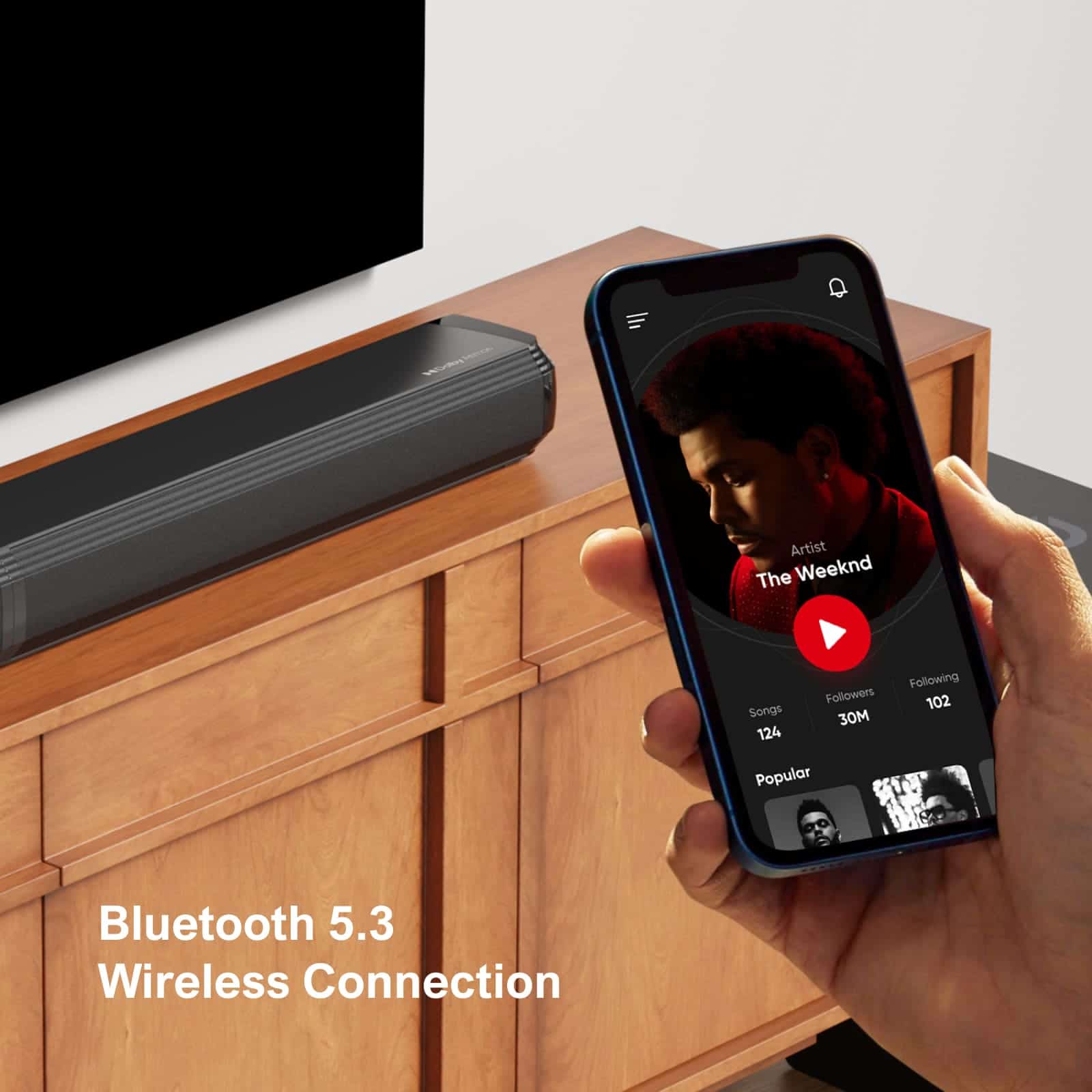 High-quality Bluetooth 5.3 wireless speaker on wooden cabinet, with a person's hand holding a smartphone showing music app, showcasing modern audio technology.
