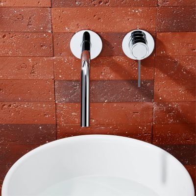 Sleek modern bathroom faucet with chrome finish mounted on red brick wall, above a white ceramic sink, showcasing contemporary bathroom design and fixtures.
