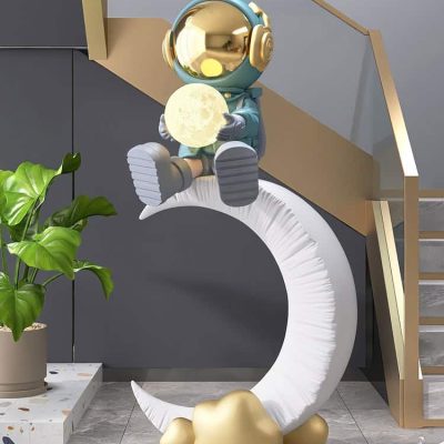 Shiny astronaut figurine sitting on a crescent moon sculpture holding the moon, modern interior decor with a green plant, stylish staircase, and artistic design elements in Monalisa Store.