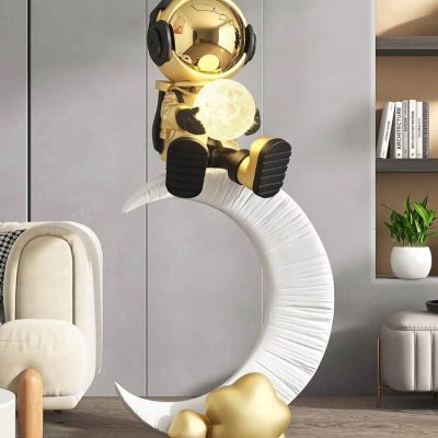 Shiny gold astronaut figurine holding a moon-shaped light, sitting on a modern white crescent moon sculpture in a stylish home interior setting.