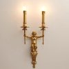 Ornate gold wall sconce featuring a cherubic figure holding dual candle holders, ideal for elegant interior lighting decoration. Luxurious vintage style lighting fixture for sophisticated spaces.