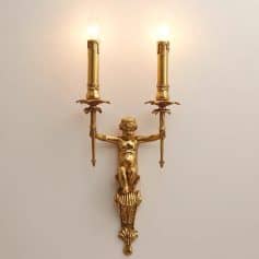 Ornate gold wall sconce featuring a cherubic figure holding dual candle holders, ideal for elegant interior lighting decoration. Luxurious vintage style lighting fixture for sophisticated spaces.