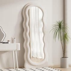 Luxurious freeform white upholstered floor mirror with wavy edges in modern minimalist bedroom decor, reflecting natural light from sheer curtains, complemented by a potted palm and skincare products.