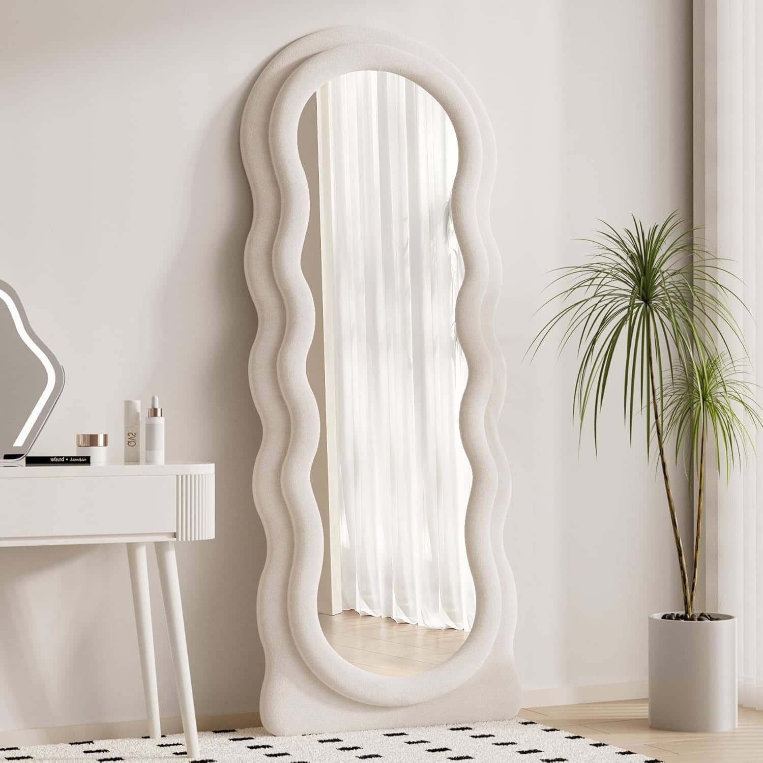 Luxurious freeform white upholstered floor mirror with wavy edges in modern minimalist bedroom decor, reflecting natural light from sheer curtains, complemented by a potted palm and skincare products.
