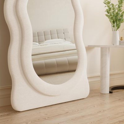 Elegant beige upholstered bed with a tufted headboard, reflected in a stylish, sculptural full-length mirror in a modern, minimalist bedroom setting.